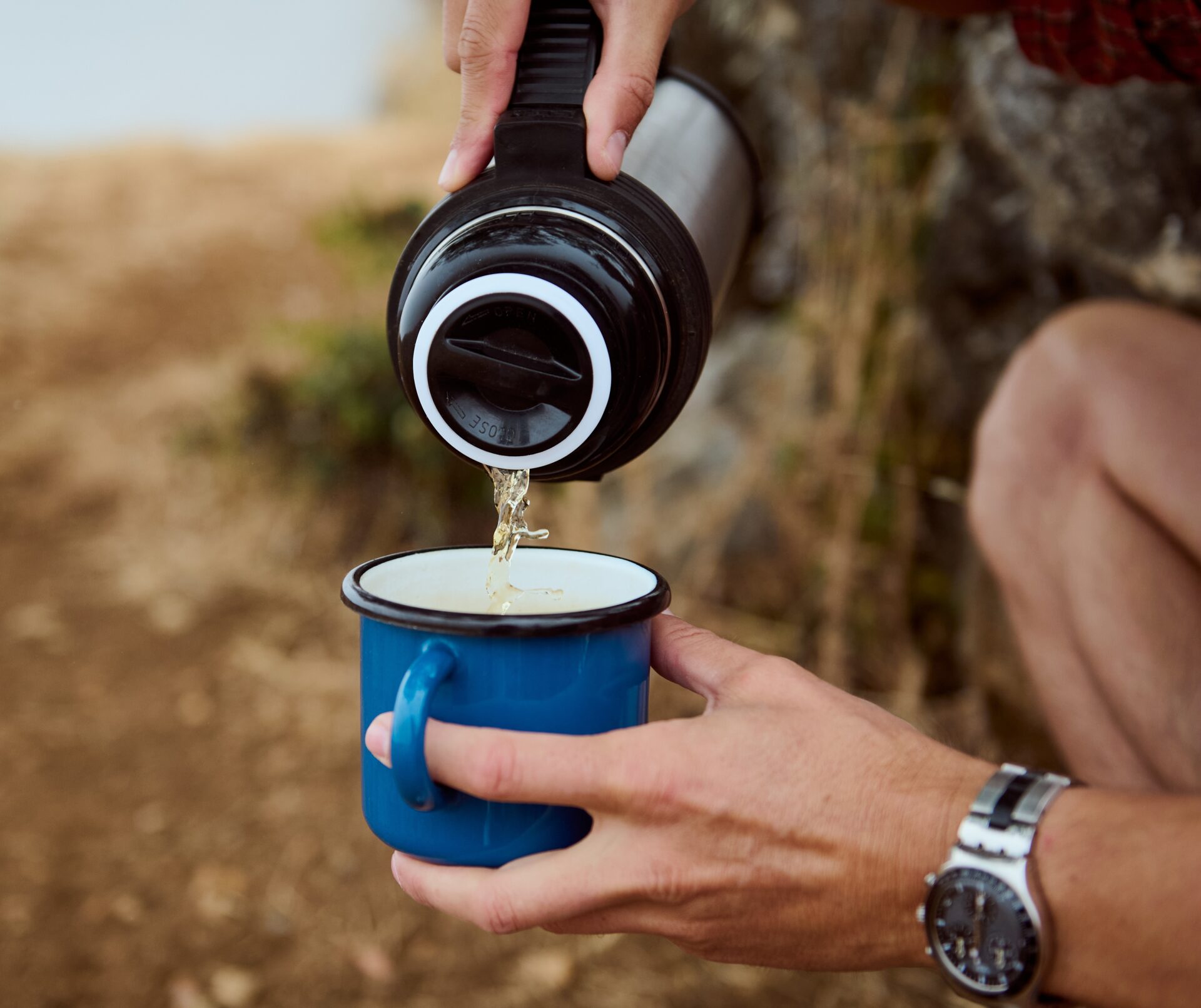 Kuuma kuppi pitkään – miten valita oikea termospullo? Close-up of hands pouring hot beverage into blue camping mug outdoors